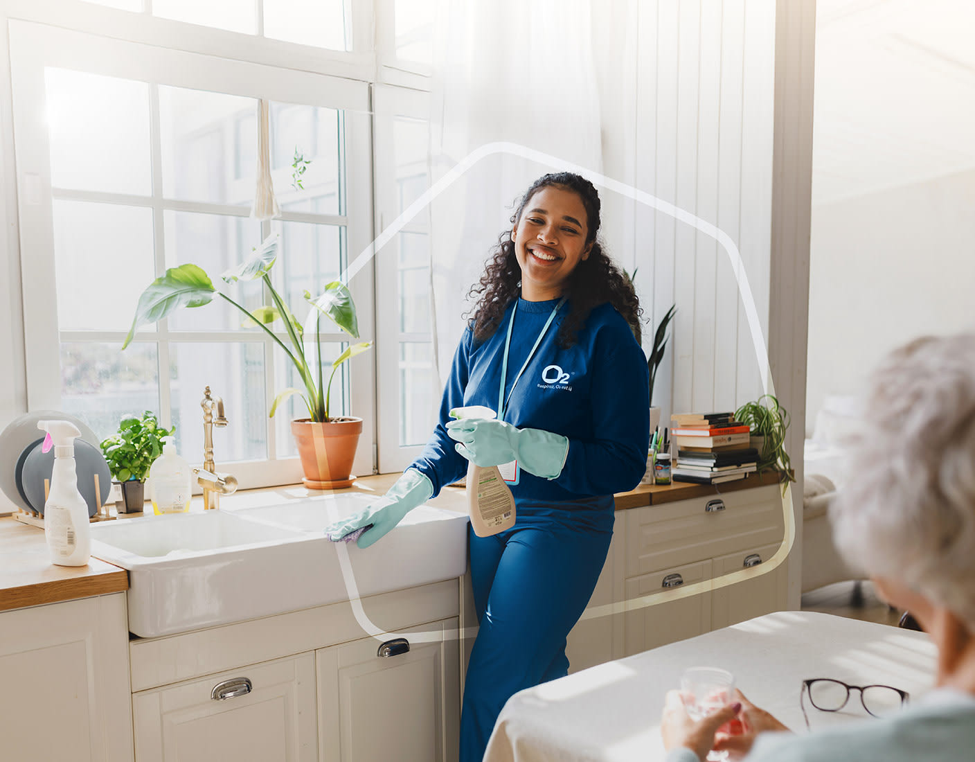 A woman in blue O2 uniform is smiling and diligently cleaning the kitchen sink.
